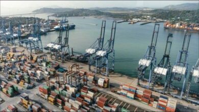 A large container ship at a busy global shipping port with stacks of containers and cranes.