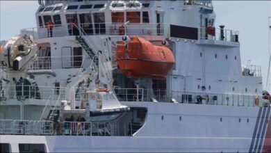 A partially submerged blue fishing boat capsizes in rough waters with rescue personnel nearby.