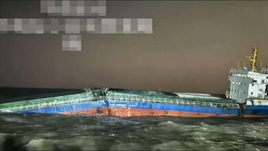 A large cargo ship emits heavy smoke from its midsection, with a rescue vessel positioned nearby.