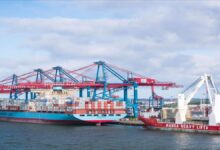 A large Maersk container ship is docked at a bustling port terminal, surrounded by stacked cargo containers.