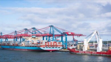 A large Maersk container ship is docked at a bustling port terminal, surrounded by stacked cargo containers.