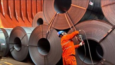 Large steel coils stacked on a dock next to a bulk carrier ship at a port.