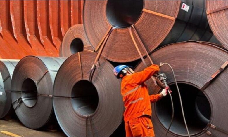 Large steel coils stacked on a dock next to a bulk carrier ship at a port.