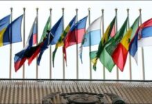 Numerous national flags fly against a blue sky, symbolizing IMO ship registration and maritime nations.