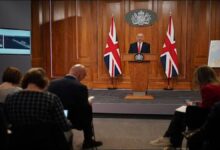 A grey naval warship sails at sea with an aircraft flying overhead.