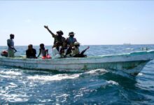 Armed Somali pirates in a small skiff approach a large cargo ship at sea.