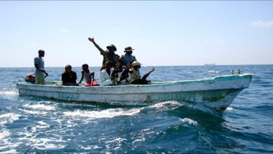 Armed Somali pirates in a small skiff approach a large cargo ship at sea.