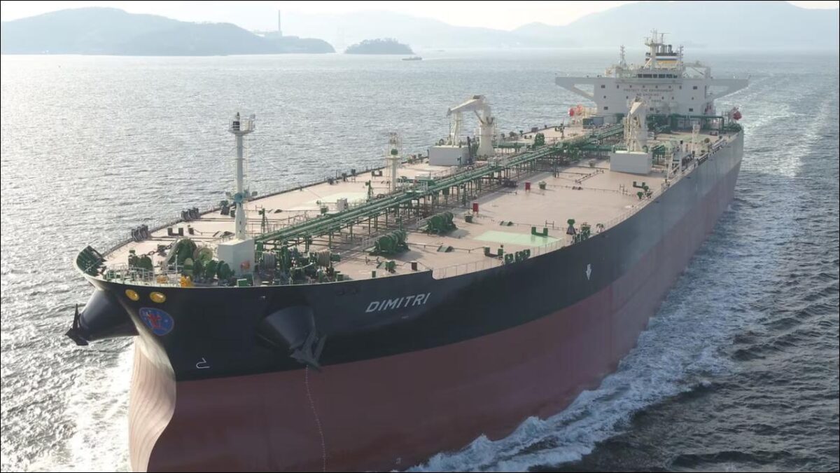 A large modern vessel, possibly a crude oil carrier, is guided by tugboats at a shipyard.