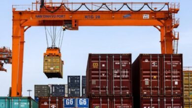 A large container ship packed with colorful containers maneuvers through a busy port.
