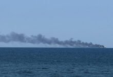A cargo ship docked near port infrastructure with smoke rising from a recent attack in Odesa.