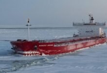 An ice-class cargo vessel navigates through thick ice in an Arctic region with snow-capped mountains.