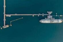 A large container ship docked at a modern deep-water port with cranes and stacked containers.