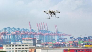 A drone hovering near a large commercial container ship docked in a busy international port.